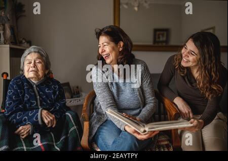 Frauen Generation mit alten kranken Großmutter im Rollstuhl sitzen und lächelnde Tochter und Enkelin suchen ein Fotoalbum. Stockfoto