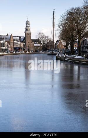 Winterstadtbild von Lemmer, Niederlande mit reformierten Kirchen und Geschäften am Kai mit einem Schiff, das im Eis von Het Dok, blauem Himmel und Sonne gefroren ist Stockfoto