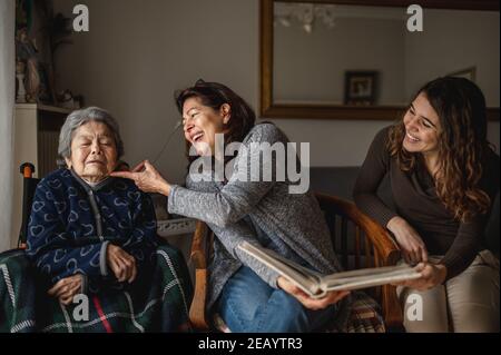 Frauen Generation mit alten kranken Großmutter im Rollstuhl sitzen und lächelnde Tochter und Enkelin suchen ein Fotoalbum. Stockfoto