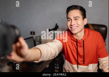 Junger lächelnder Mann, der die Kamera und andere professionelle Geräte aufsetzt, um ein Video am Schreibtisch zu machen. Vlog, Konzept für freiberufliche Arbeit. Stockfoto