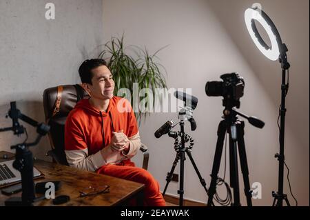 Junger Mann, der im Studio ein Video von sich selbst im Gespräch mit der Kamera aufnimmt. Arbeitsplatz mit Mikrofon, Stativen, Kamera und Ringleuchten. Vlog-Konzept Stockfoto