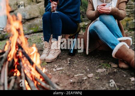 Unkenntliche Frauen halten ein Glas Rotwein am Lagerfeuer und wärmen sich neben einem Feuer. Stockfoto