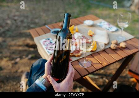 Nahaufnahme nicht erkennbare Hand hält eine Flasche Wein. Tischset mit Vorspeisen und leckeres Essen im Hintergrund. Weinprobe und Picknick-Konzept. Stockfoto