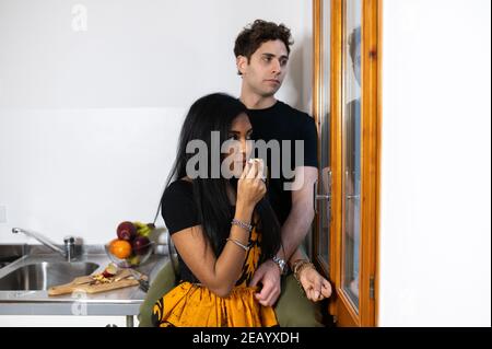 Junges Paar zu Hause essen Apfel in der Küche. Indische Frau mit traditionellen Kleid lächelt zu kaukasischen Mann auf der Theke sitzend und schaut aus der Stockfoto