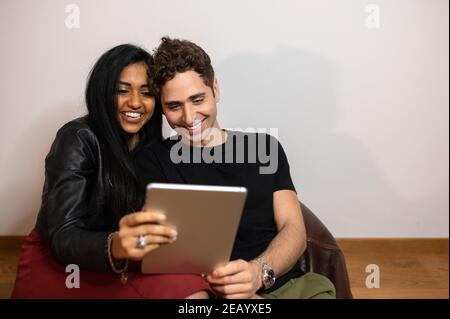 Junge multi-ethnische Paar Spaß zu Hause sitzen auf einem Sessel, während Sie einen Film auf Tablet. Stockfoto