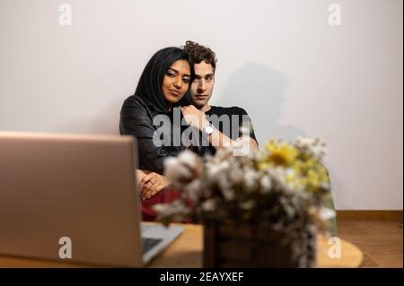 Junges multiethnisches Paar sitzt auf einem Sessel und schaut sich einen Film am Computer zu Hause an. Frau auf Männerbeinen Stockfoto