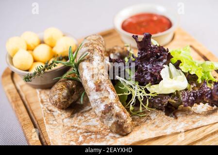 Portion schmackhafter gegrillter hausgemachter Würstchen mit gekochter Kartoffel, Salat und herzhafter Tomatensauce auf Holzbrett Stockfoto