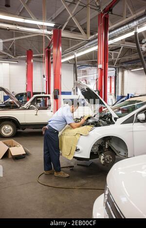Der hispanische Mechaniker lehnt sich an ein Auto an, während er daran arbeitet Der Motorraum in einer Autowerkstatt Stockfoto