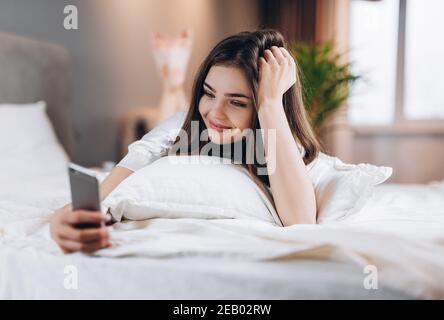Junge Frau, die mit einem Lächeln auf ihrem Gesicht ins Telefon schaut. Morgens oder abends im Bett mit einem Telefon. Brunette Scrollen das Futter während im Bett liegen. Frau Stockfoto