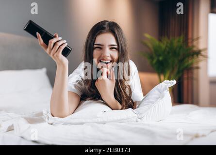 Frau in weißer Seide Pyjamas schaut TV im Schlafzimmer auf dem Bett. Weibliche Modell mit einer tv-Fernbedienung in den Händen liegend auf dem Bett. Morgen oder Stockfoto