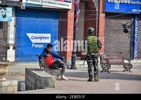 Srinagar, Indien. Februar 2021, 11th. Ein Jugendlicher benutzt sein Handy als paramilitärischer Trooper, der während des Stillstands durch einen geschlossenen Markt patrouilliert.IN Kaschmir WIRD ZUM 37th. Todestag des Gründers der Jammu und der Kashmir Liberation Front (JKLF), Mohammad Maqbool Bhat, ein Shutdown beobachtet. Der an diesem Tag im Jahr 1984 im Tihar-Gefängnis in Neu Delhi aufgehängt wurde. Kredit: SOPA Images Limited/Alamy Live Nachrichten Stockfoto