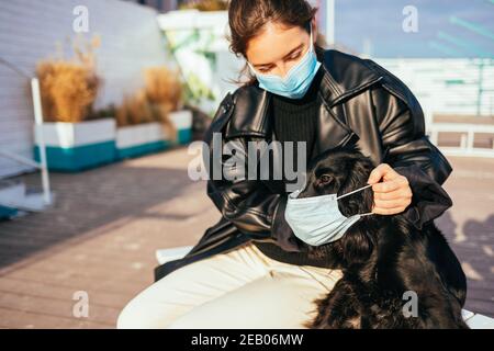Glückliche junge Frau trägt medizinische Maske setzt auf Schutzmaske auf ihren Hund zu, während draußen am sonnigen Tag sitzen. Stockfoto