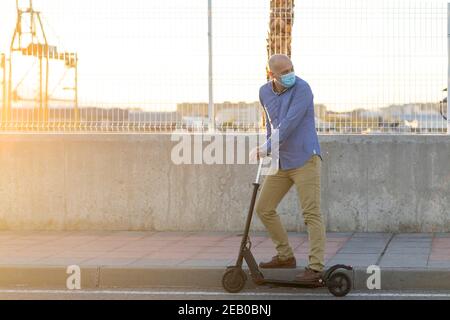 Reifer Mann trägt eine Maske nutzt einen Elektroroller in der Stadt. Er trägt blaues Hemd und ockerfarbene Hosen. Es gibt Licht bei Sonnenuntergang. Horizontales Foto. Suchen Stockfoto
