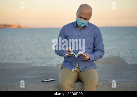 Mann liest Buch auf der Straße, trägt Maske. Das Meer ist im Hintergrund zu sehen. Er hat ein Mobiltelefon. Horizontales Foto mit Platz zum Einfügen von Text. Stockfoto