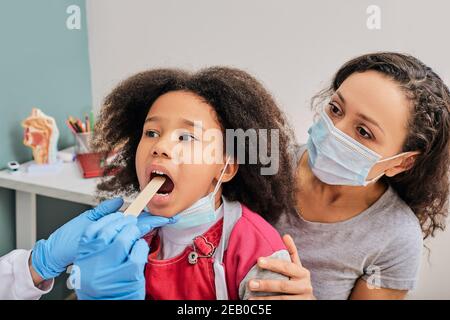 Doktor benutzt Inspektionsspachtel, um die Kehle eines afroamerikanischen Mädchens zu untersuchen. Kleines Mädchen mit ihrer Mutter beim Kinderarzt-Termin Stockfoto