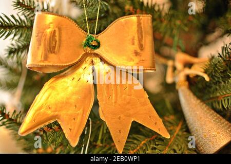 Goldene Weihnachtsdekoration, die an einem Ast im grünen Baum hängt, in Form eines Bogens (Knoten) mit weichem Bokeh. Nahaufnahme, Vorderansicht Stockfoto