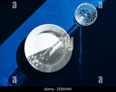 Tischeinstellung mit leerer weißer Platte und Kristallglas isoliert auf dunkelblauem Hintergrund, dunklen Schatten und Sonnenlicht, das durch das Weinglas reflektiert wird Stockfoto