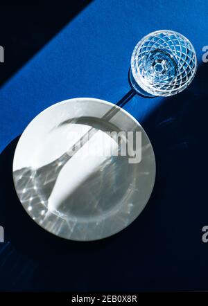 Tischeinstellung mit leerer weißer Platte und Kristallglas isoliert auf dunkelblauem Hintergrund, dunklen Schatten und Sonnenlicht, das durch das Weinglas reflektiert wird Stockfoto