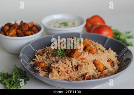 Tomatenreis serviert mit Raita und gebratenen Hähnchenbissen ohne Knochen. Aufnahme auf weißem Hintergrund. Serviert auf einer grauen Schüssel Stockfoto