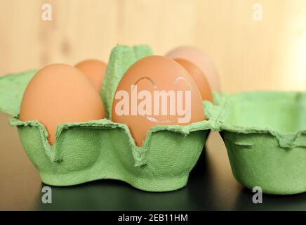 Gruppe von braunen Hühnereiern in einer grünen offenen Box auf natürlichem Holzhintergrund. Das nächste Ei ist mit weißem Smiley bemalt. Vorderansicht, Nahaufnahme Stockfoto