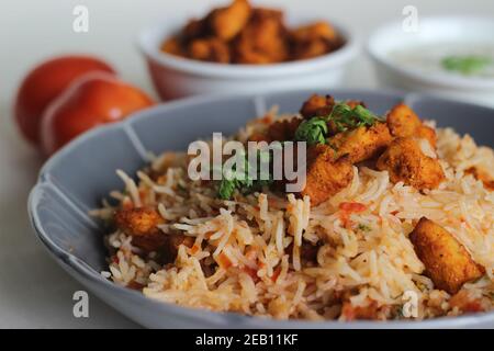 Tomatenreis serviert mit Raita und gebratenen Hähnchenbissen ohne Knochen. Aufnahme auf weißem Hintergrund. Serviert auf einer grauen Schüssel Stockfoto