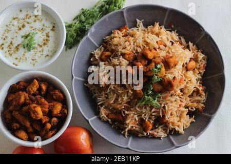 Tomatenreis serviert mit Raita und gebratenen Hähnchenbissen ohne Knochen. Aufnahme auf weißem Hintergrund. Serviert auf einer grauen Schüssel Stockfoto