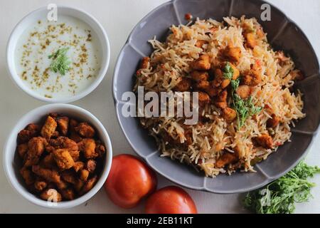 Tomatenreis serviert mit Raita und gebratenen Hähnchenbissen ohne Knochen. Aufnahme auf weißem Hintergrund. Serviert auf einer grauen Schüssel Stockfoto