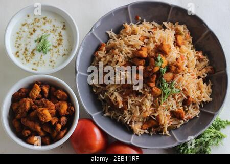 Tomatenreis serviert mit Raita und gebratenen Hähnchenbissen ohne Knochen. Aufnahme auf weißem Hintergrund. Serviert auf einer grauen Schüssel Stockfoto