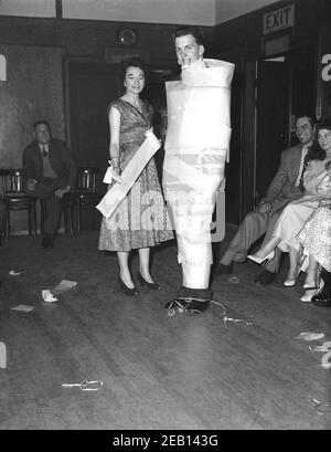 1950s, historisch, bei einer Geburtstagsparty in einem Veranstaltungsraum eines Hotels, ein großer junger Mann neben einer Frau stehend Hals bis Fuß in braunem Papier gewickelt und hält ein kleines Geschenk in seinem Mund, ein Partyspiel dieser Zeit kein Zweifel, England, Großbritannien. Die neben ihm stehende Dame hält eine Papierrolle, die vermutlich selbst die Tat vollbracht hat. Stockfoto