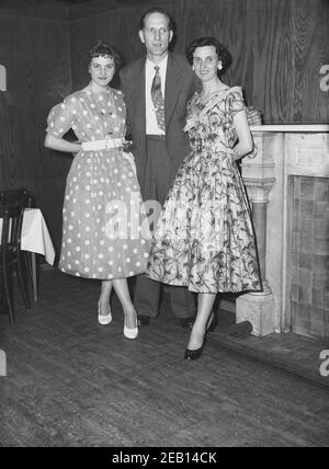 1950s, historische, junge Dame in einem gefleckten oder gepunkteten Kleid modisch in dieser Zeit steht für ein Bild mit einem Paar bei ihrem Geburtstag Pary in einem Hotel Veranstaltungsraum, England, Großbritannien. Stockfoto