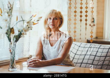 Portrait von elegant attraktive blonde Kaukasierin eine gealterte Frau mit stilvollen Haaren in weißem T-Shirt sitzt an einem Esstisch in der Nähe des Fensters, Blick auf Stockfoto