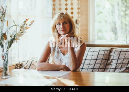 Portrait von elegant attraktive blonde Kaukasierin eine gealterte Frau mit stilvollen Haaren in weißem T-Shirt sitzt an einem Esstisch in der Nähe des Fensters, Blick auf Stockfoto
