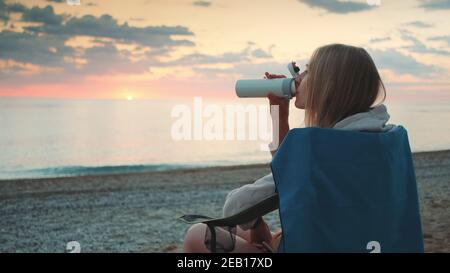 Junge Frau trinkt aus Thermoskannen und sitzt auf Campingstühlen am Strand. Profilansicht. Stockfoto