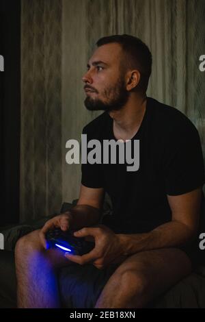 Junger schöner Gamer Mann mit Bart in schwarzem T-Shirt spielen Videospiel mit Joystick mit einer Konzetration auf Smart Face Ernsthaft denken in den Abendgarden Stockfoto
