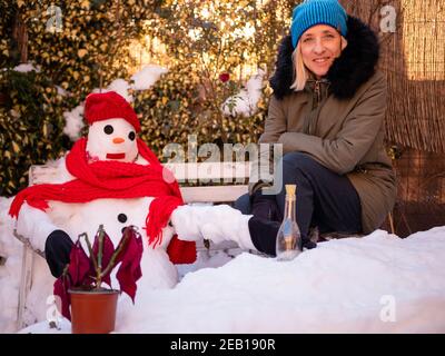 Blonde Frau sitzt neben einem Schneemann, in einem Park mitten im Winter mit allem Schnee Stockfoto