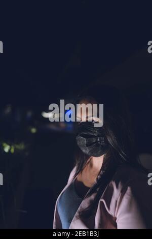 Frau trägt Gesicht Sicherheit schwarze Maske ihr Gesicht beleuchtet trägt eine Maske gegen Coronavirus, Straße ohne Menschen im Hintergrund Stockfoto