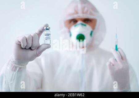 Gesundheitshelfer in Schutzanzug und Maske hält eine Injektionsspritze und Impfstoff. Biologische Gefahr. Stockfoto