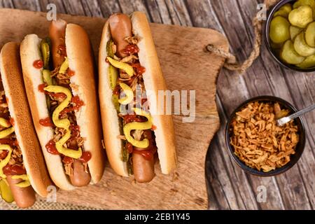 Hausgemachte Hotdogs mit Würstchen und Brötchen mit eingehaltener Gurke, getrockneten Zwiebeln, Senf und Ketchup neben Schüsseln mit Zutaten gekrönt Stockfoto