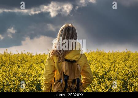 Sturm und Regen kommen. Wanderfrau, die im Rapsfeld steht und den wolkigen Himmel anschaut. Tourist trägt gelbe wasserdichte Jacke Stockfoto