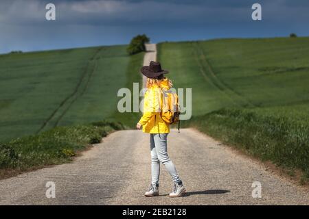 Regen kommt. Frau, die auf der Straße wandert und den wolkenlosen Himmel anschaut. Backpacker trägt Hut und gelbe wasserdichte Jacke Stockfoto