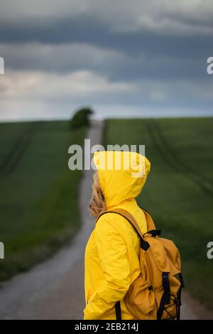 Regen kommt. Frau, die auf der Straße wandert und den wolkenlosen Himmel anschaut. Backpacker trägt gelbe wasserdichte Jacke mit Kapuze Stockfoto
