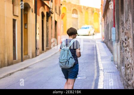 Ein Touristenmädchen mit gestreiftem Rucksack geht eine sonnendurchflutete europäische Straße hinunter. Keine Teilfläche sichtbar. Reisen durch Spanien, Barcelona. Hoher pH-Wert Stockfoto