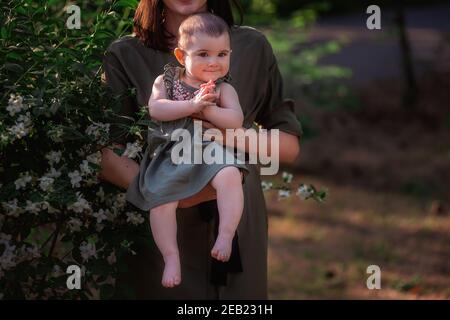 Nahaufnahme eines glücklichen kleinen Babys, das ihre Hände in die Arme einer schönen Mutter klatscht. Junge Frau geht mit ihrer Tochter in einem grünen Park in der Nähe von Th Stockfoto