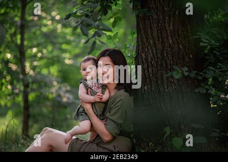 Eine gesunde junge Mutter hält ein Kleinkind in den Armen. Eine glückliche Familie sitzt auf dem grünen Gras, unter einem hohen Baum, spielt, umarmt, genießt einen Spaziergang in der Stockfoto