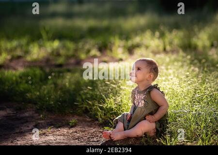 Ein kleines niedliches Baby mit kurzen Haaren, in einem grünen Kleid sitzt auf dem Rasen im Gras in der Nähe des Weges, schaut in den Himmel in der Sonne. Sie lächelt sie an Stockfoto