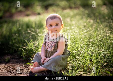 Ein kleines niedliches Baby mit kurzen Haaren, in einem grünen Kleid sitzt auf dem Rasen im Gras in der Nähe des Weges, schaut in den Himmel in der Sonne. Sie lächelt sie an Stockfoto