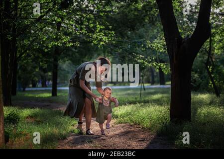 Das kleine Mädchen macht die ersten Schritte, eine junge Mutter hält ein Kind bei den Händen, versichert, hilft, den Weg des Parks inmitten grüner Bäume zu gehen Stockfoto