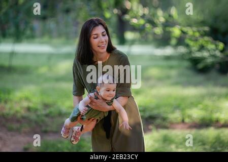 Junge braune Mutter schüttelt ihr Baby in den Armen im Park unter den Bäumen, grünes Gras. Frau hält kleines Mädchen in den Armen, spielt, umarmt Kind Stockfoto