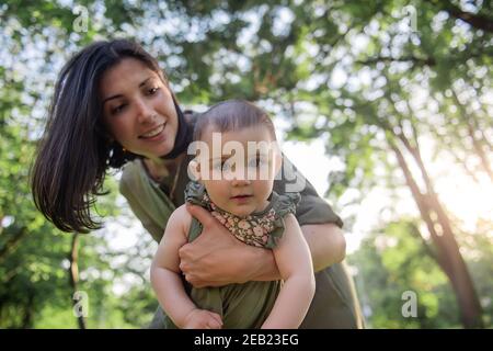 Junge braune Mutter schüttelt ihr Baby in den Armen im Park unter den Bäumen, grünes Gras. Frau hält kleines Mädchen in den Armen, spielt, umarmt Kind Stockfoto