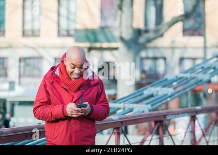 Kahler lateinischer Mann in einem roten Mantel mit seinem Telefon Beim Gehen in einer Straße Stockfoto